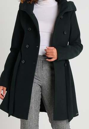 Woman wearing a dark green double-breasted coat over a white turtleneck and gray plaid pants, holding the coat's lapel with one hand.