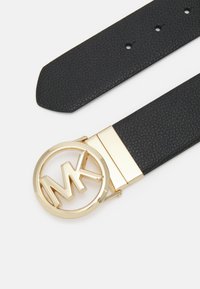 Svart texturerad läderbälte med en guldfärgad cirkulär spänne med "MK"-logotyp. Spännet har en elegant design med en polerad yta.