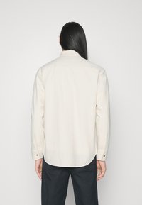 Chemise beige clair à coupe décontractée, manches longues, poignets boutonnés, col classique et ourlet arrondi. Texture lisse en coton.