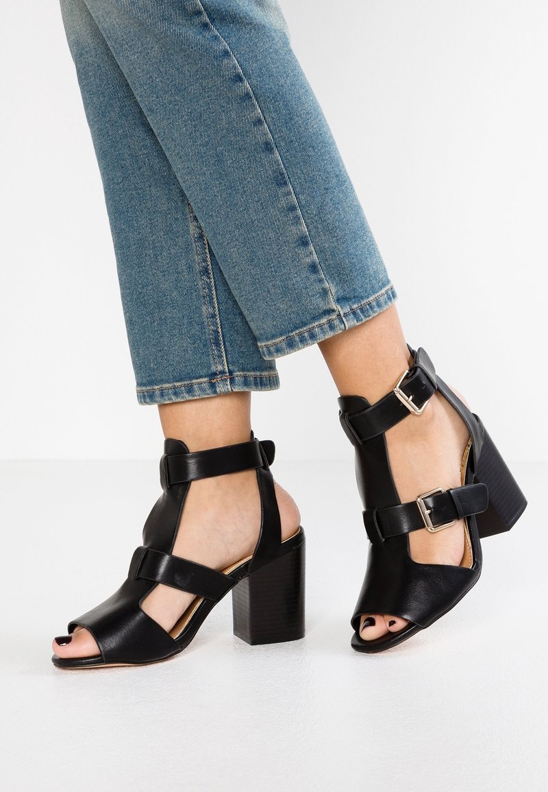 Sorte læderhøjhælede sandaler med åben tå, der har ankelremme og spænder. Design med blokhæle; glat tekstur og minimalistisk stil.