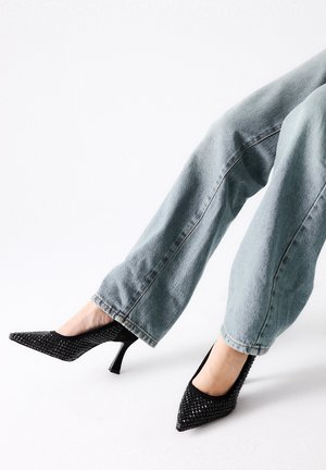 Zapatos de tacón negro con textura trenzada, punta afilada y tacón grueso, combinados con jeans de pierna ancha en azul claro.