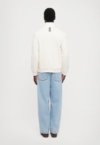 Hvit hettegenser med ribbet mansjetter og kant, med et svart vertikalt stripe-design på baksiden. Kombinert med lyseblå vide jeans.