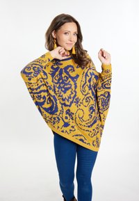 Gehaakte poncho in geel met een blauw paisleypatroon, met brede mouwen en een hoge hals. Gekleed over blauwe jeans en bruine laarzen.