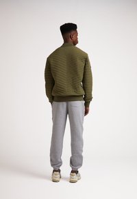Giubbotto bomber trapuntato verde con un motivo zigzag testurizzato, abbinato a pantaloni jogger grigi e sneaker, visto di spalle.