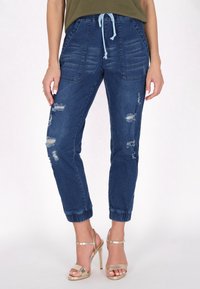 Sötétkék farmer joggerek húzózsinórral a deréknál, szakadás részletekkel, oldalsó zsebekkel és elasztikus mandzsettákkal a bokán. Arany magassarkú szandálokkal párosítva.