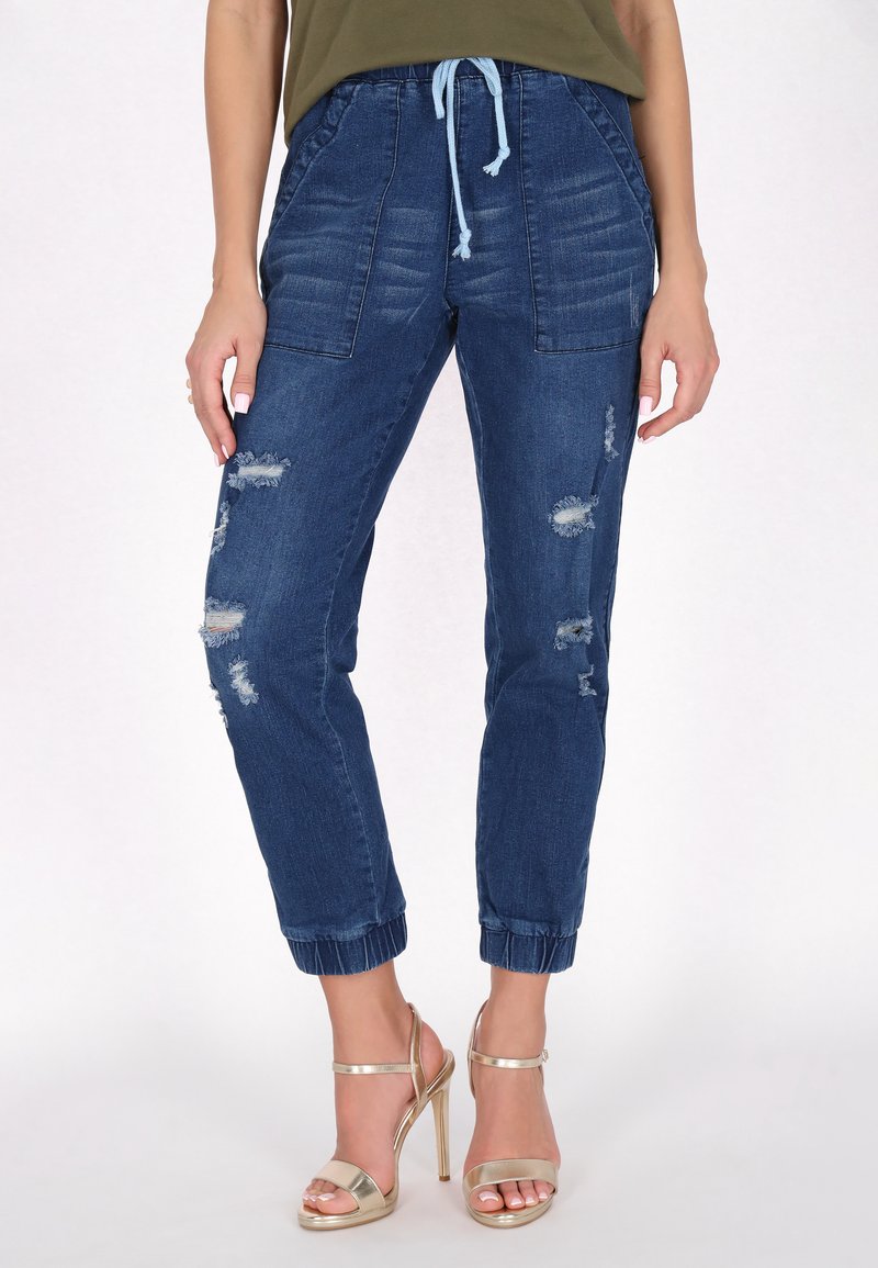 Sötétkék farmer joggerek húzózsinórral a deréknál, szakadás részletekkel, oldalsó zsebekkel és elasztikus mandzsettákkal a bokán. Arany magassarkú szandálokkal párosítva.