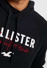 Svart huvtröja med vit och röd text. Har en känguruficka och dragsnodd i huvan, tillverkad av mjukt material.