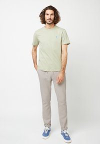 Hellgrünes Baumwoll-T-Shirt mit einem kleinen blauen Baum-Graphic, kombiniert mit beigen Herrenhosen und blauen Sneakers, vor einem schlichten Hintergrund stehend.