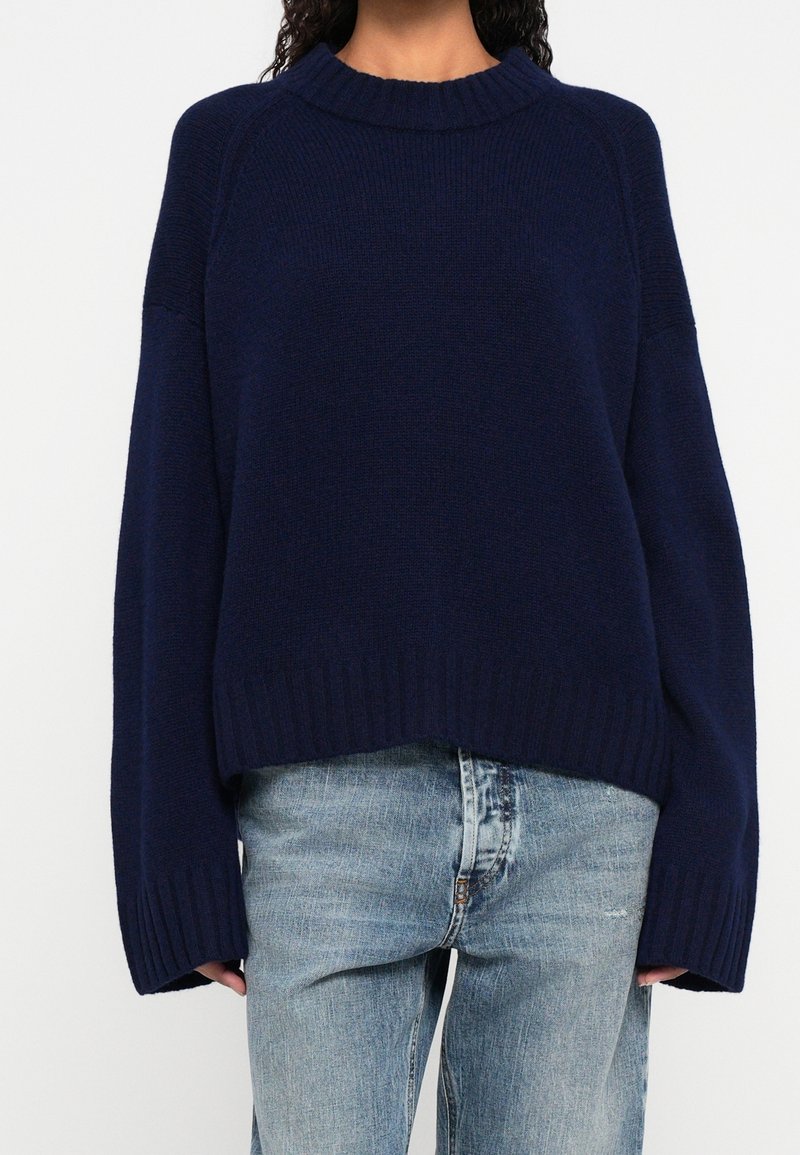 Personne portant un pull tricoté oversize bleu foncé à manches longues et un jean en denim bleu clair, devant un fond uni.