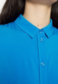 Camicetta blu brillante con colletto, dal tessuto liscio, dotata di chiusura con bottoni e con bottoni visibili. Cuciture ordinate e design classico.
