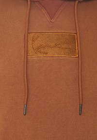 Sweat à capuche marron avec cordons assortis et un patch brodé rectangulaire avec texte en écriture cursive centré sur la poitrine.