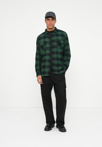 Camicia di flanella a quadri verde e nera con chiusura a bottoni, due tasche sul petto, abbinata a pantaloni cargo neri e scarpe nere.