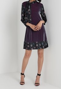 Lila Kleid mit langen Ärmeln, das einen floral gemusterten schwarzen unteren Teil und eine Halsbinde aufweist, aus glattem, poliertem Stoff.