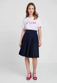 Junge Frau mit lockigem Haar, trägt ein weißes T-Shirt mit pinkem Text, einen marineblauen Faltenrock und pinke High-Heels, steht vor einem schlichten Hintergrund.