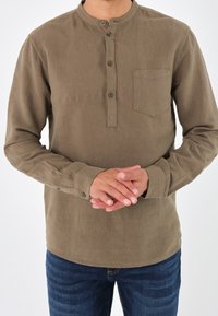 Camicetta marrone a maniche lunghe con colletto rotondo, patta frontale con bottoni, taschino sul petto e tessuto strutturato, abbinata a jeans blu.