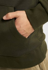 Mørk grønn hettegenser med frontlomme, som viser en strukturert stoff og ribbestrikkede mansjetter. Hånden hviler i lommen.