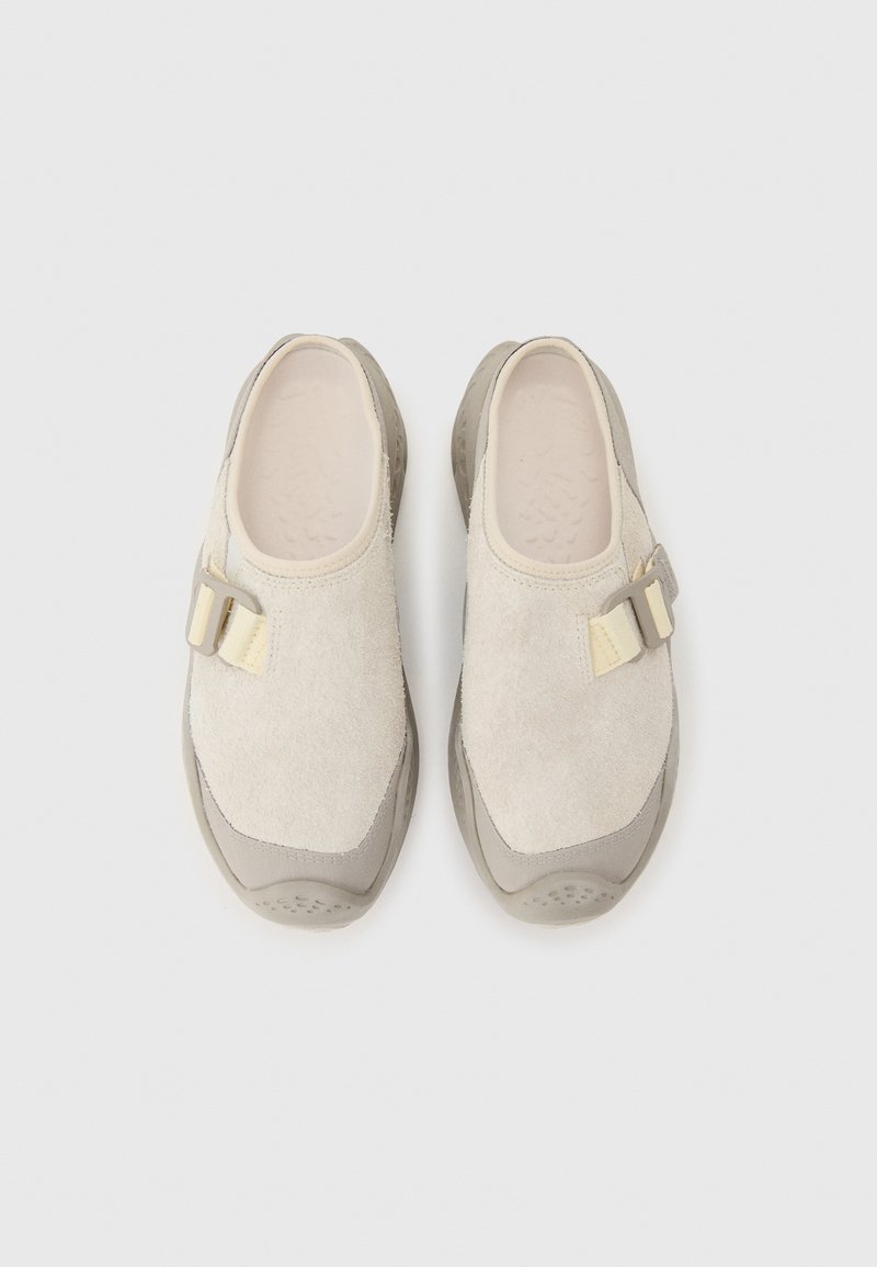 Beige suède instapsloffen met ronde neus, verstelbare zijriem en gestructureerde grijze rubberen zool, gezien van bovenaf op een lichte achtergrond.