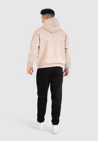 Sweat à capuche beige clair avec un grand texte blanc dans le dos, associé à un pantalon noir et des baskets blanches. Tissu doux et décontracté.