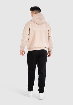 Light beige hooded sweatshirt with large white text on the back, paired with black pants and white sneakers. Soft, casual fabric.