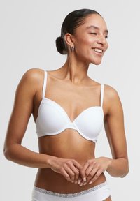 Bílý vyztužený podprsenka s vlnitým krajkovým lemem, nastavitelné ramínka a hladký povrch. Uložena na modelce, zdůrazňující design.