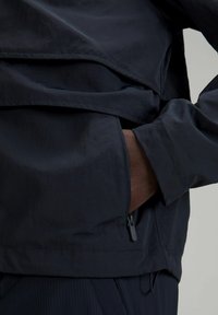 Námořnický větrovka s texturovaným povrchem, se zapínáním na půl zip a boční kapsou, zdůrazňující lehký, vrstvený materiál.
