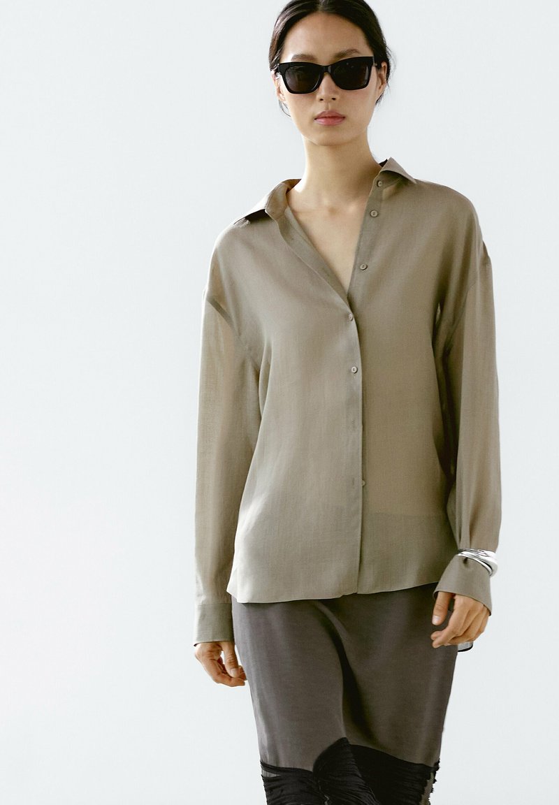 Olijfgroene button-up shirt van lichtgewicht stof met een kraag en lange mouwen, gecombineerd met een donkergrijze rok met textuurelementen.