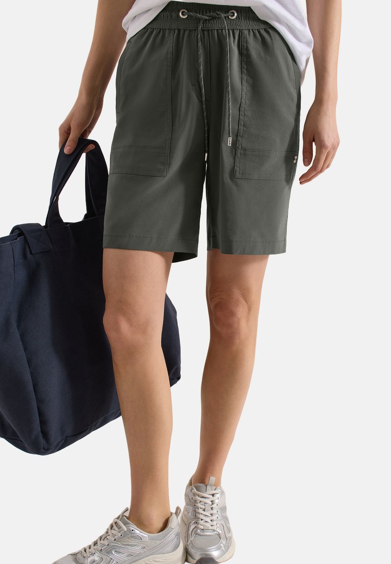 Personne portant un short vert foncé à cordon, des baskets blanches, un haut blanc, tenant un grand sac cabas bleu foncé à ses côtés.