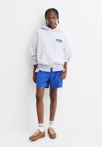Grå hætte sweatshirt med blå tekst, lagdelt over en blårandet skjorte, parret med blå shorts og brune ruskinds sneakers.