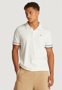 Camisa polo blanca de mangas cortas con rayas rojas y azul marino en los puños. Cuello clásico y pequeño logo en el lado izquierdo del pecho.