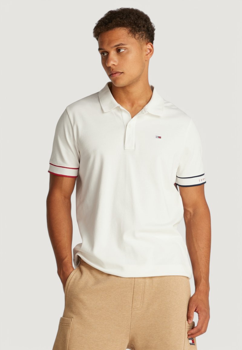 Camisa polo blanca de mangas cortas con rayas rojas y azul marino en los puños. Cuello clásico y pequeño logo en el lado izquierdo del pecho.