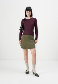 Un haut violet à rayures et manches longues associé à une jupe vert olive. Les accessoires comprennent un petit sac à main noir et des chaussettes beiges avec des chaussures pointues noires.