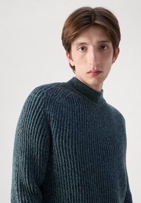 Gestrickter Pullover in dunklem Petrolblau mit gerippter Textur, hohem Kragen und leicht lockerer Passform. Bietet reiche Details und einen gemütlichen Look.