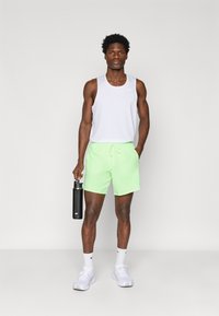 Αθλητής που φοράει λευκό αμάνικο μπλουζάκι, νεον πράσινα σορτς, λευκά αθλητικά παπούτσια και κάλτσες, κρατώντας ένα μαύρο μπουκάλι νερού της Nike, στέκεται μπροστά σε απλή φόντο.