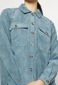 Giacca camicia in corduroy blu chiaro con colletto, chiusura frontale a bottoni, due tasche sul petto e texture a righe verticali.