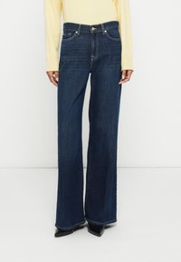 Široke kavbojke iz temnega jeansa z visokim pasom, sprednjimi žepi in zapiranjem na gumb. Kombinirane s črnimi čevlji z ostrim sprednjim delom.