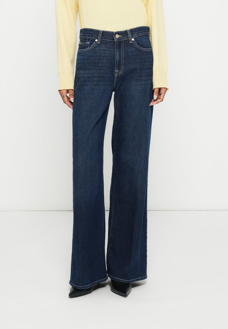 Široke kavbojke iz temnega jeansa z visokim pasom, sprednjimi žepi in zapiranjem na gumb. Kombinirane s črnimi čevlji z ostrim sprednjim delom.