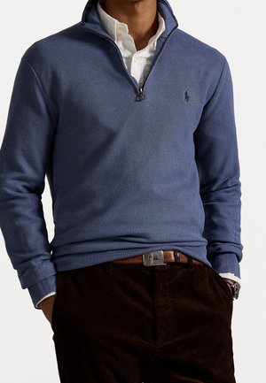 Homme portant un pull bleu à demi-fermeture éclair sur une chemise blanche à boutons, ceinture marron et pantalon foncé, les mains dans les poches, sur un fond uni.