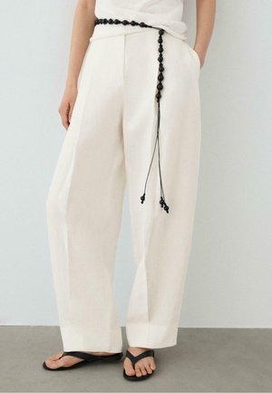 Personne portant un pantalon blanc ample avec une ceinture noire nouée et des tongs noires, debout avec une main dans la poche devant un fond uni.