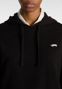Hoodie noir avec cordons, arborant un petit motif de skateboard blanc sur la poitrine. Superposé à une chemise de couleur claire.