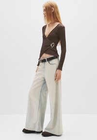 Brun, struktureret indpakningstop med detaljer i sølvring, parret med lyseblå, vide jeans og sort bælte, sat mod en ensfarvet baggrund.