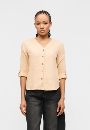 Kvinne med flettet hår iført en beige knappet bluse med oppbrettede ermer og falmede svarte jeans, stående foran en ensfarget bakgrunn.