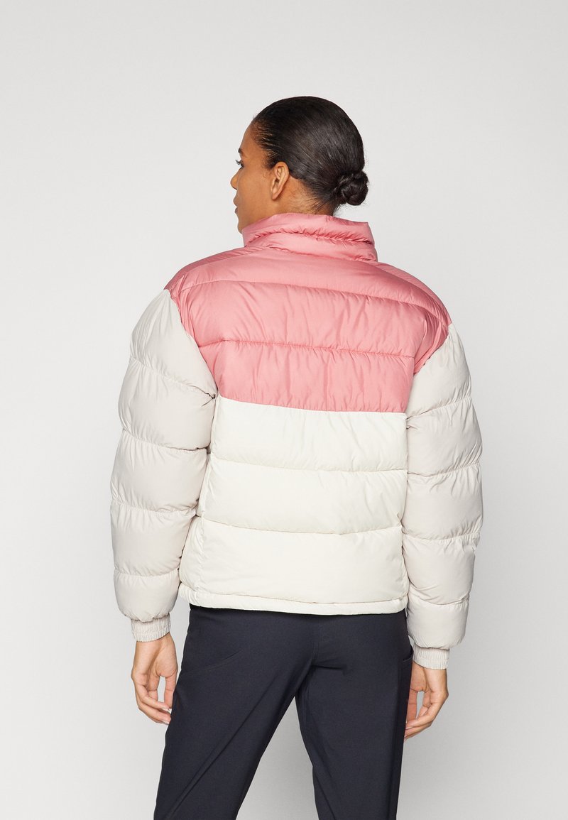 Mujer de piel oscura y cabello recogido en un moño, vistiendo una chaqueta acolchada en tonos rosa y blanco con bloques de color y pantalones negros, vista desde atrás contra un fondo simple.