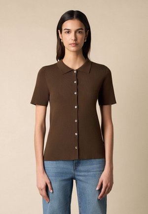 Vrouw met steil donker haar, gekleed in een bruine korte mouwen poloshirt met knopen en lichtblauwe spijkerbroek, staand tegen een beige achtergrond.