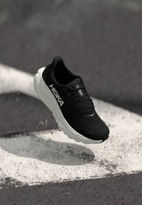 Baskets de course noires avec tige en mesh, semelle intermédiaire blanche et lacets détaillés. Logo affiché de manière bien visible sur le côté. Design légèrement surélevé.