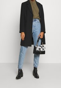 Musta pitkä takki oliivinvihreän paidan päällä, yhdistetty vaaleansinisiin farkkuihin ja mustiin nilkkureihin. Pieni käsilaukku, jossa valkoinen ja musta kuvio.