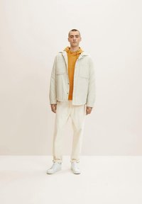 Chaqueta ligera de color crema sobre una sudadera naranja, combinada con pantalones beige claros y zapatillas blancas. Tela suave, cierre de botón, dos bolsillos.