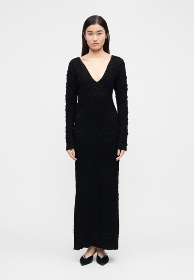Frau trägt ein strukturiertes schwarzes Maxikleid mit langen Ärmeln und tiefem V-Ausschnitt sowie schwarze spitze Schuhe, steht vor einem schlichten Hintergrund.