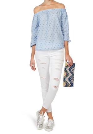Blusa azzurro chiaro off-the-shoulder con piccoli motivi blu scuro, jeans skinny bianchi strappati, sneakers argentate e una clutch multicolore.