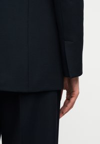 Δεξί χέρι χαλαρό δίπλα σε άτομο που φοράει σκούρο, κομψό σακάκι κοστουμιού και matching παντελόνι, πάνω σε απλό φωτεινό φόντο.