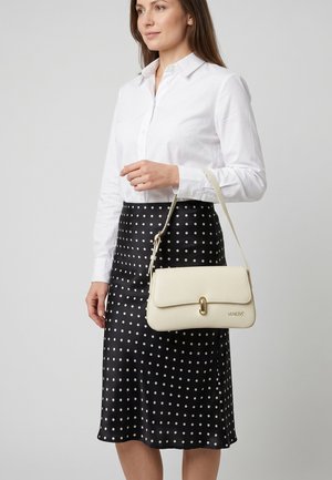Vrouw draagt een wit overhemd met knopen en een zwarte polkadot rok, houdt een crèmekleurige Venezia handtas met gouden gesp aan het handvat vast.